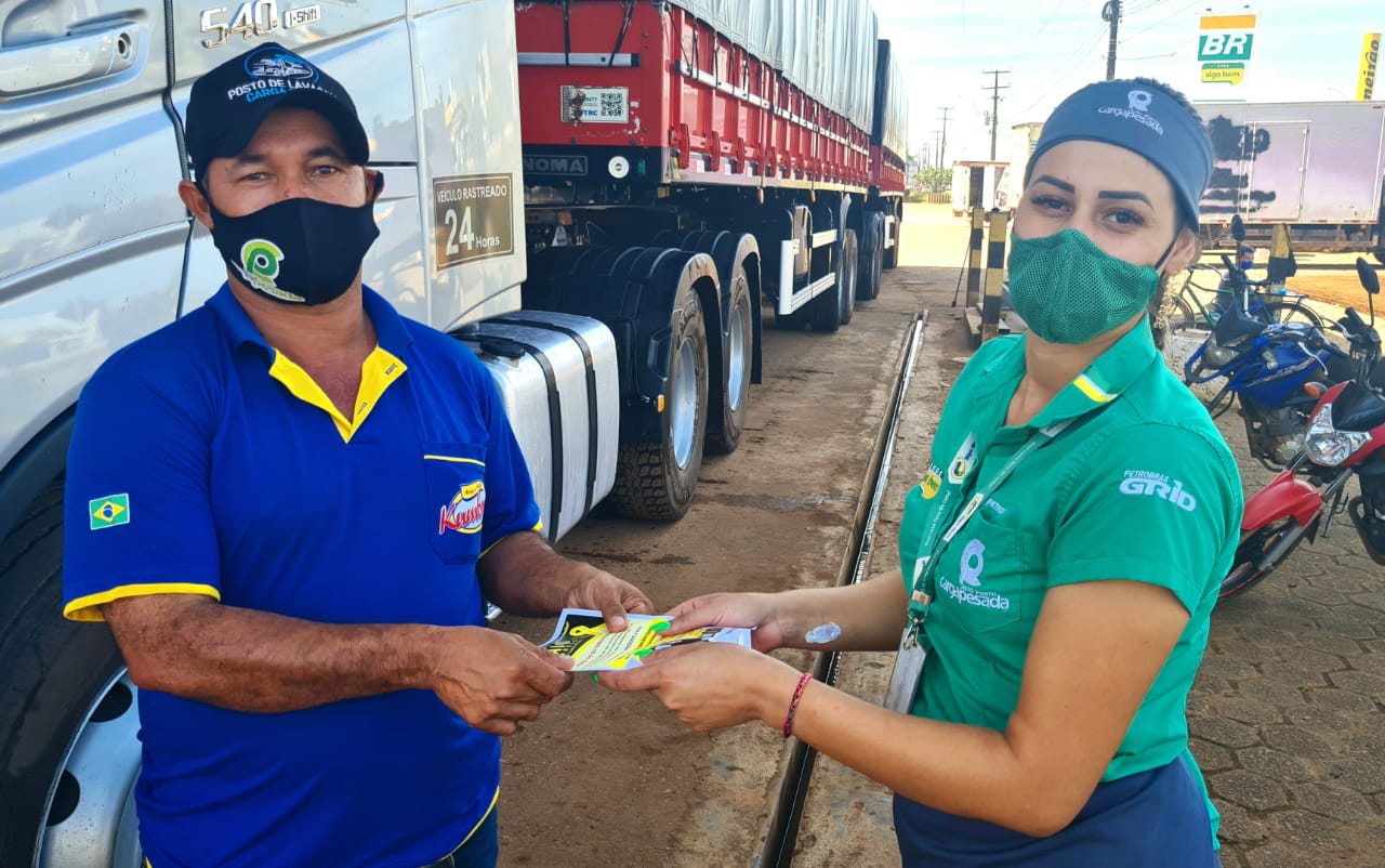 trabalhadores, com máscara, exibem panfletos de prevenção de acidentes com caminhões ao fundo
