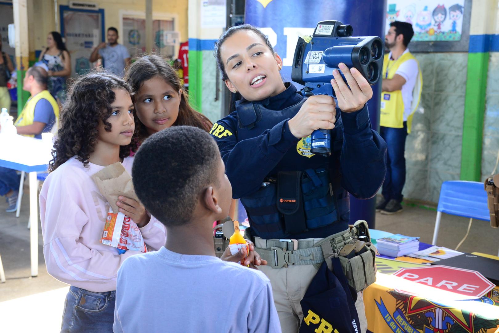 A imagem mostra a policial Rodoviária Federal mostrando um equipamento a crianças.