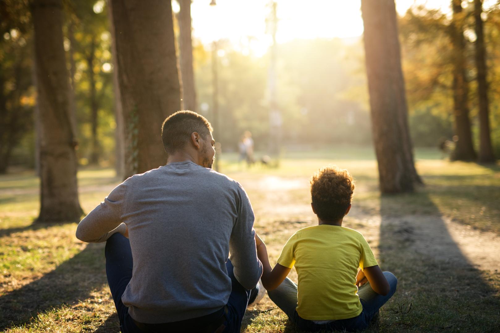 A imagem mostra pai e filho sentados no chão de uma praça