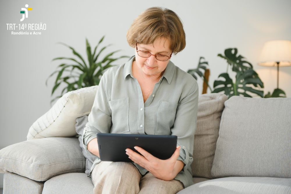 Um senhora sentada em um sofá ao centro da imagem manuseia um tablet atenta.