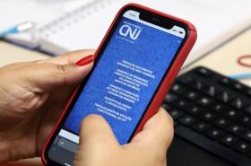Uma pessoa segura um celular com capa vermelha exibindo na tela a Revista Eletrônica do CNJ, tendo ao fundo um teclado e um caderno sobre a mesa.